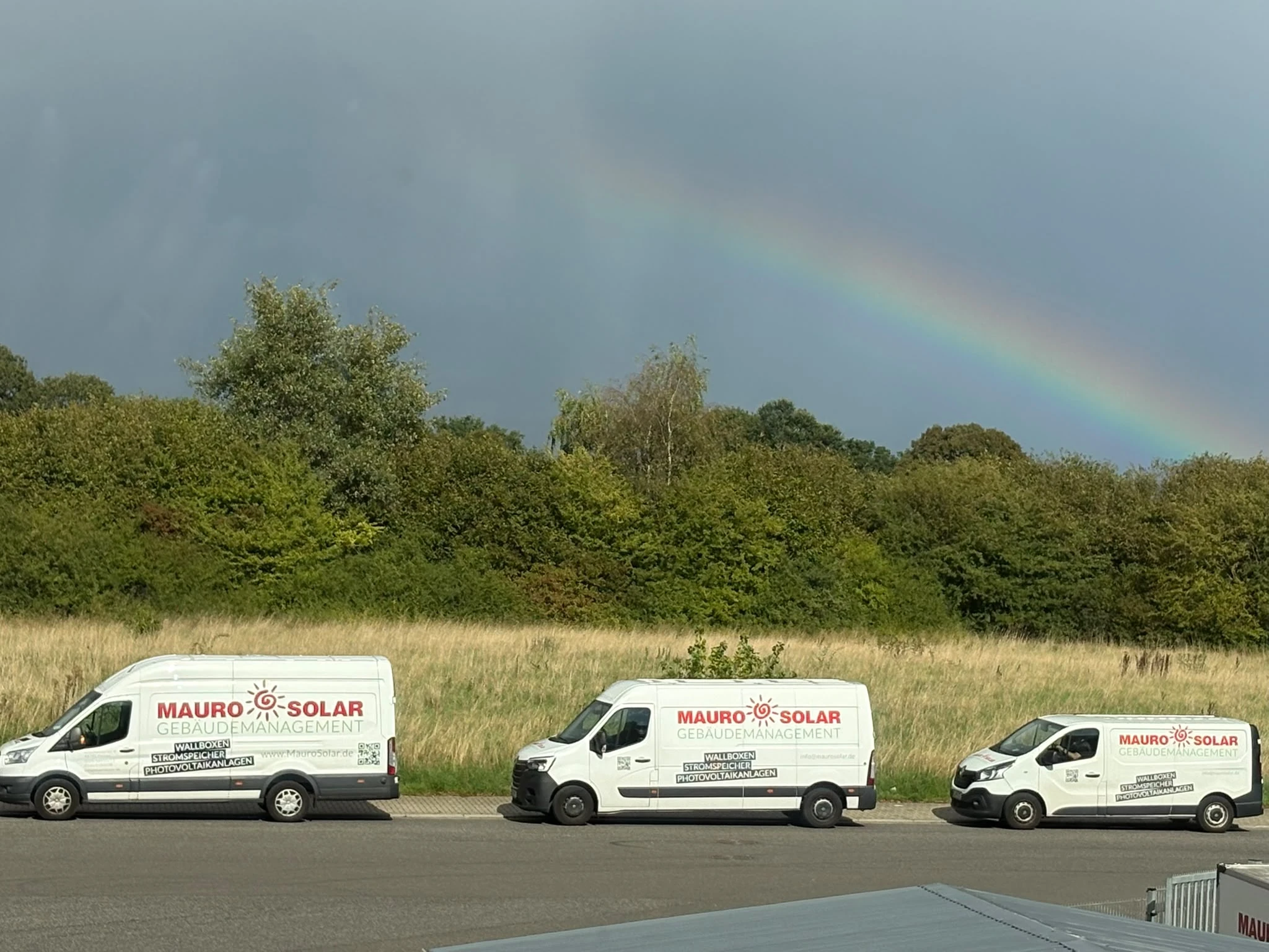 Drei Mauro-Solar-Firmenfahrzeuge vor einer Wiese mit Bäumen und einem Regenbogen.